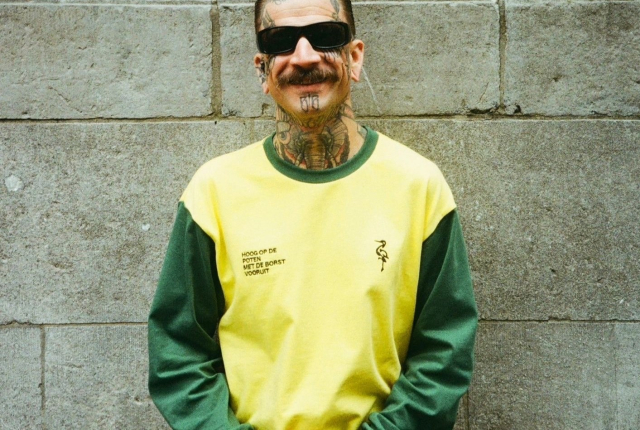 Man met tattoos poseert met een glimlach in de nieuwe collectie van Groen Gele Aderen.
