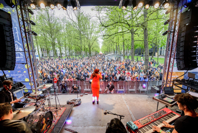 Zangeres op het podium op het Lange Voorhout tijdens The Life I Live. Ze is van achter gefotografeerd waardoor je het drukke publiek ziet.
