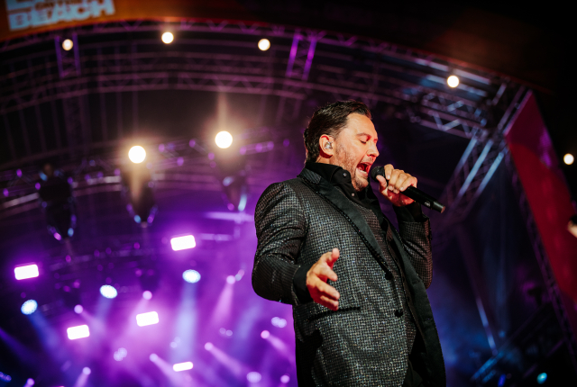 Een zanger in een glanzend kostuum zingt intens op een podium met gekleurde lichten.