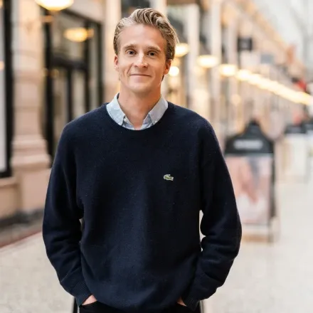 Een man in een donkerblauwe trui en overhemd staat glimlachend op straat.
