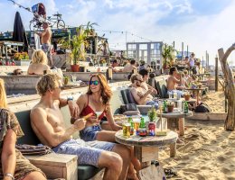 Een gezellig druk terras met mensen in badkleding met hun voeten in het zand bij strandtent Indigo op Scheveningen in Den Haag Een gezellig druk terras met mensen in badkleding met hun voeten in het zand bij strandtent Indigo op Scheveningen in Den Haag
