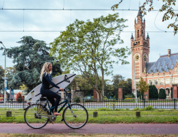 Zomer in Den Haag. Meisje met surfplank onder haar arm fiets langs het Vredespaleis richting Scheveningen Zomer in Den Haag. Meisje met surfplank onder haar arm fiets langs het Vredespaleis richting Scheveningen