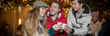 Een meisje en twee jongens proosten met warme chocomel in een sfeervol verlichte houten kraam tijdens de Royal Christmas Fair op Lange Voorhout